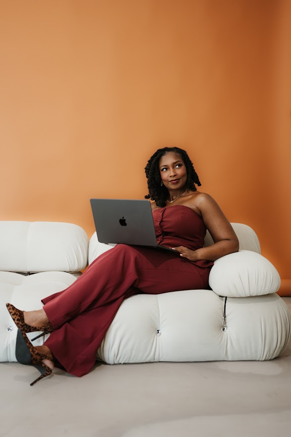 Kierra Dotson seated on a cream sofa in a burgundy set, working on a laptop